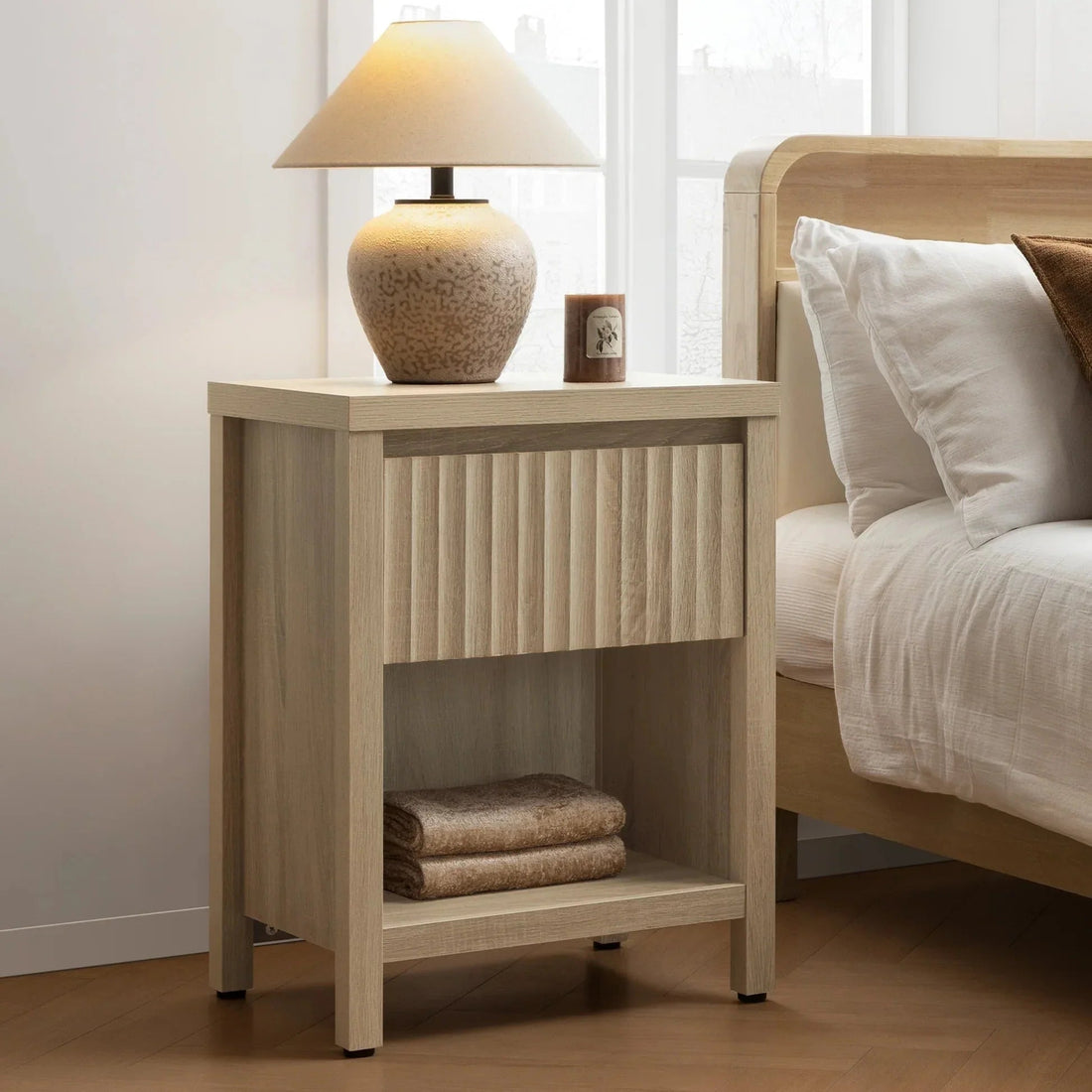 Modern wood nightstand with lamp, candle, and folded towels beside a bed