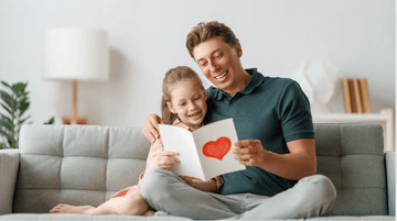 Smiling father and daughter reading a handmade heart card on a gray sofa in a bright living room
