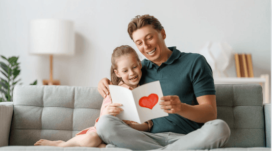 Smiling father and daughter reading a handmade heart card on a gray sofa in a bright living room