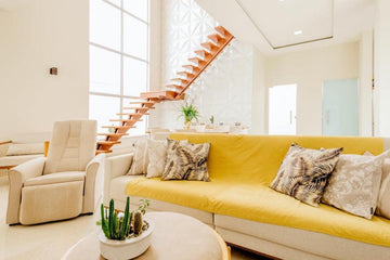 Modern living room with yellow sofa, patterned pillows, indoor plants, and wooden stairs