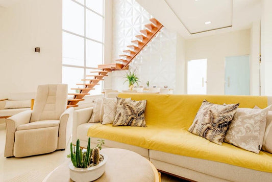 Modern living room with yellow sofa, patterned pillows, indoor plants, and wooden stairs