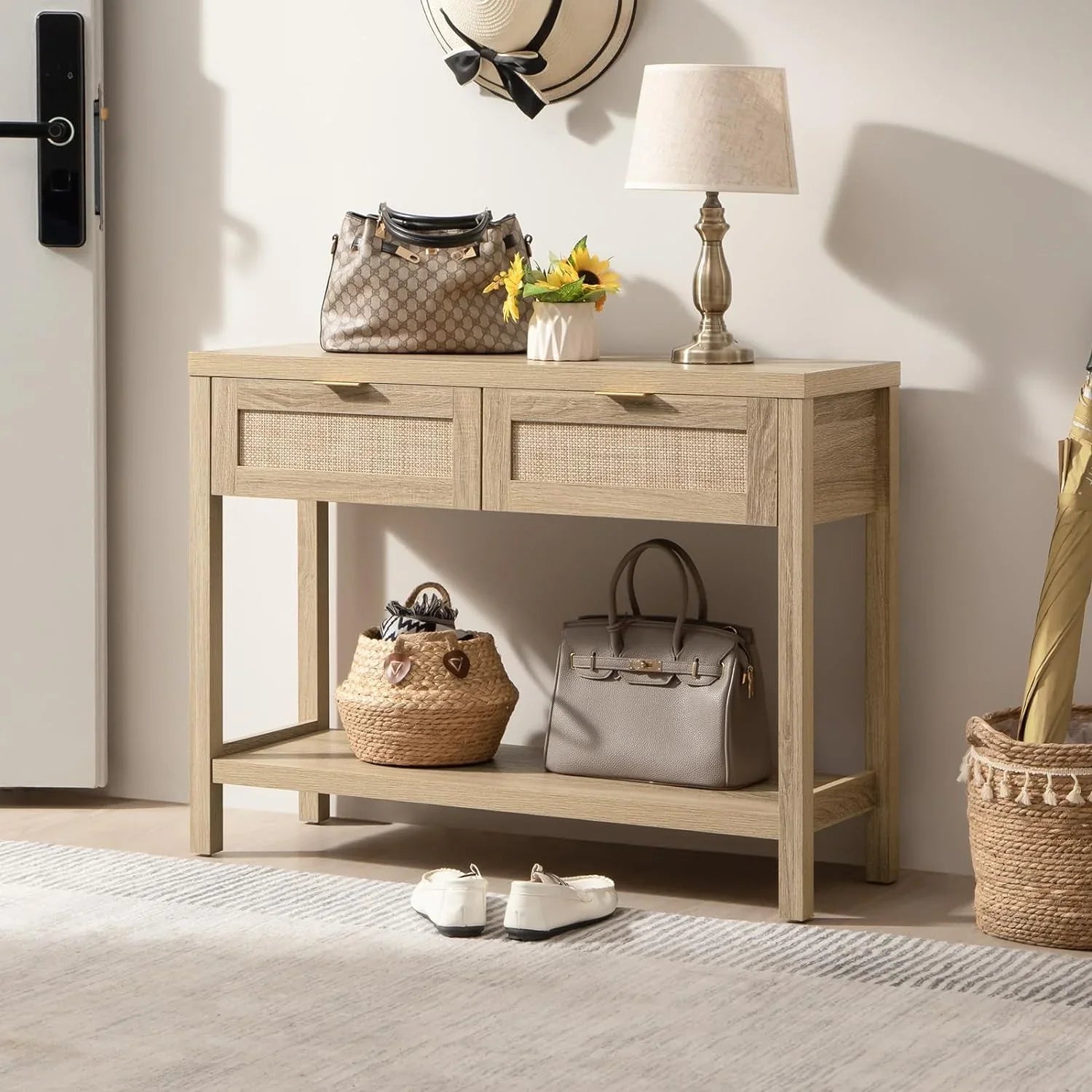 A dark wood console table with woven rattan panels on the drawers, gold handles, and a lower shelf, decorated with a vase, books, and a lamp.