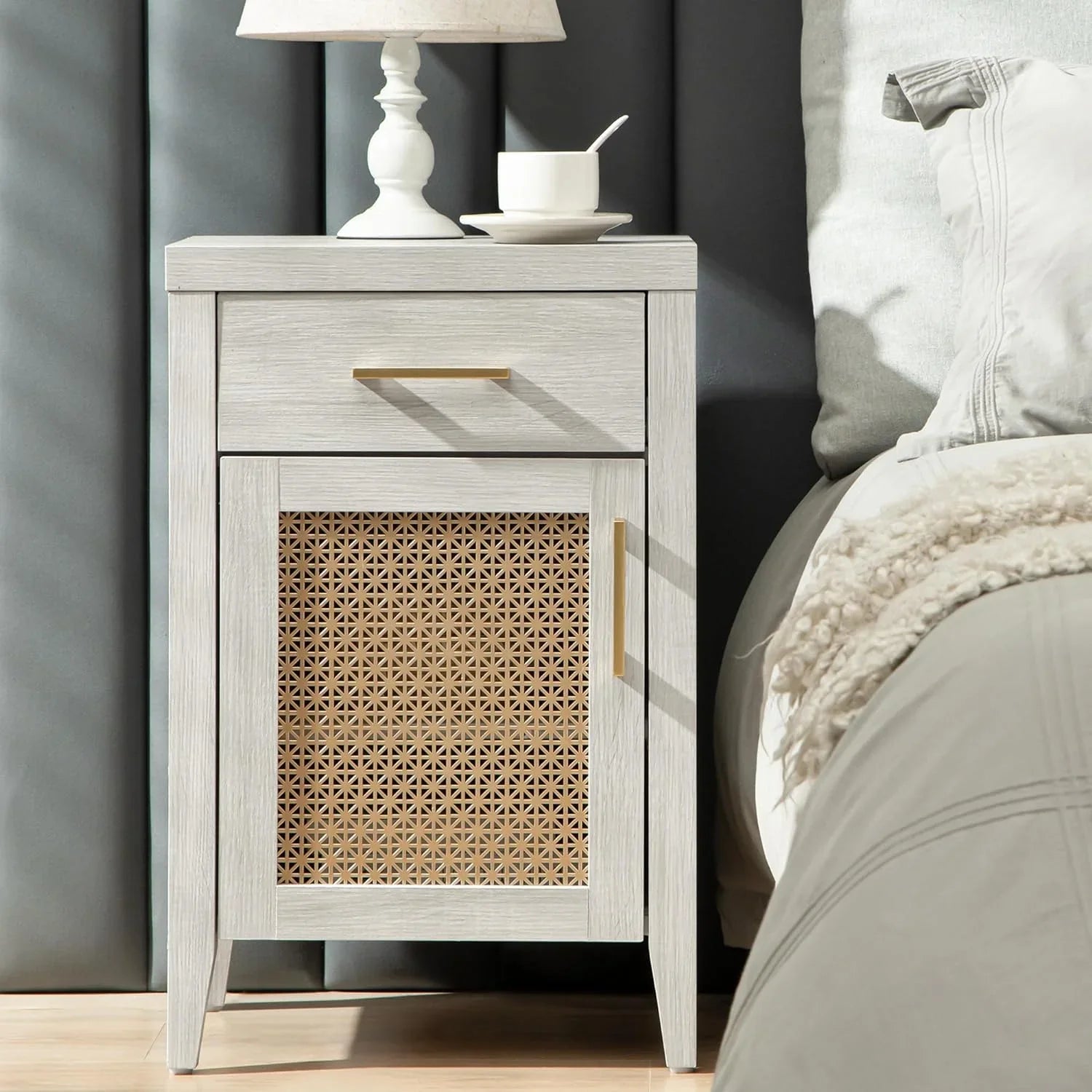 Light wood nightstand with rattan door, gold handles, lamp, and cup beside bed