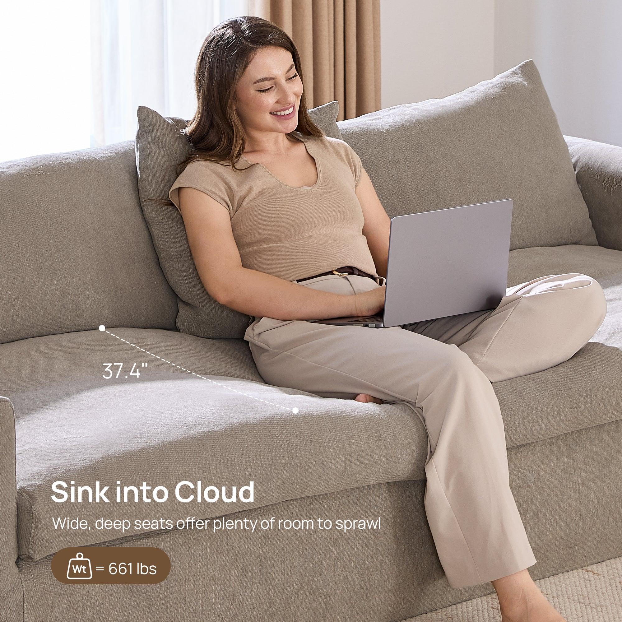 A woman reclining on a white sofa with a laptop, in a bright living room with large windows, wooden flooring, and a beige rug.