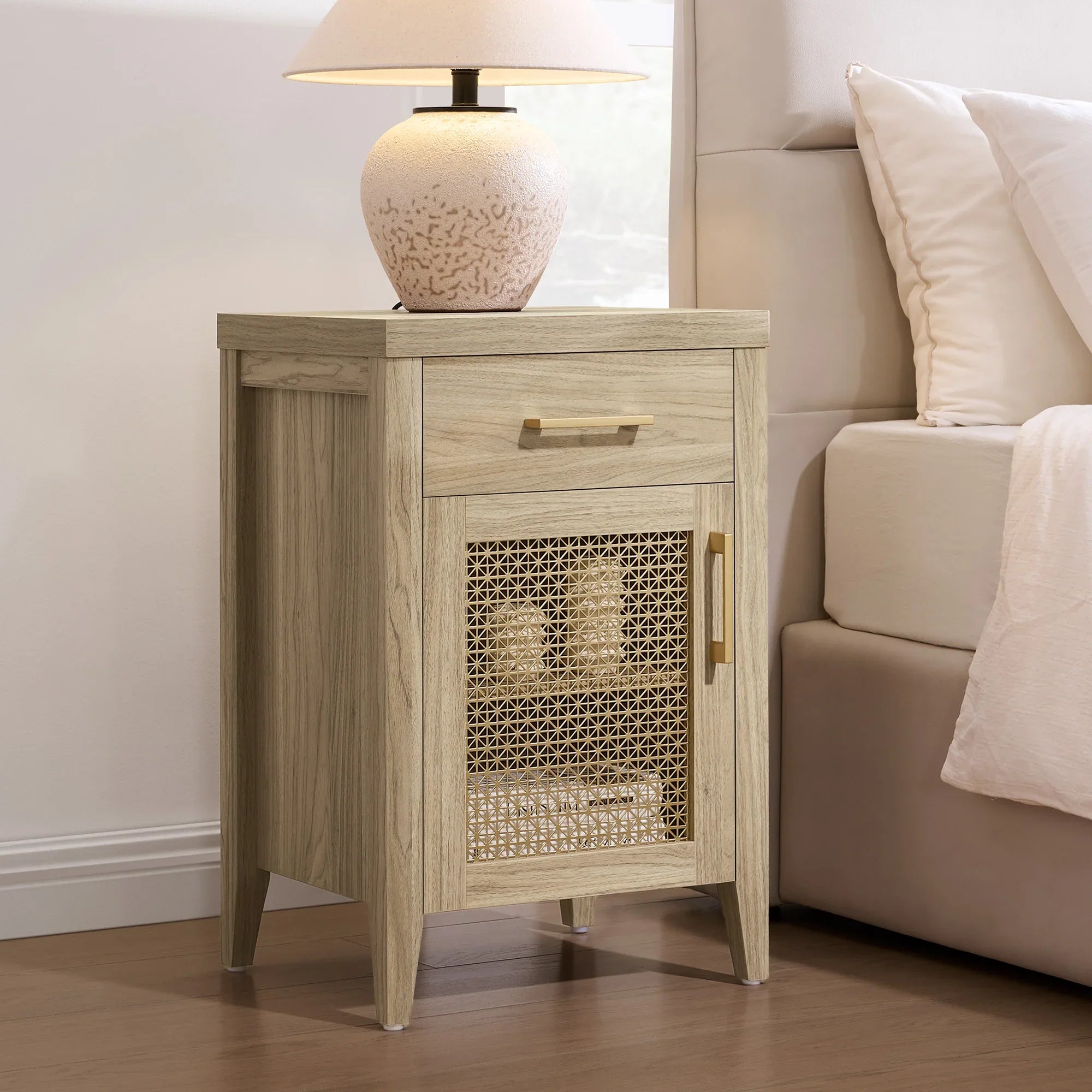 Wood nightstand with rattan door, gold handles, and a ceramic lamp beside a bed.
